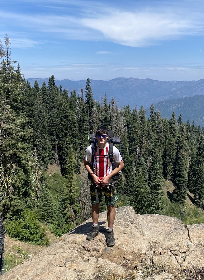 Jasper Burgard poses while hiking in the mountains. A clear, blue sky and mountains in the background.